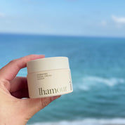 A hand holding a cream-colored jar of lhamour Hydrating Facial Cream against a blurred background of the blue ocean and sky